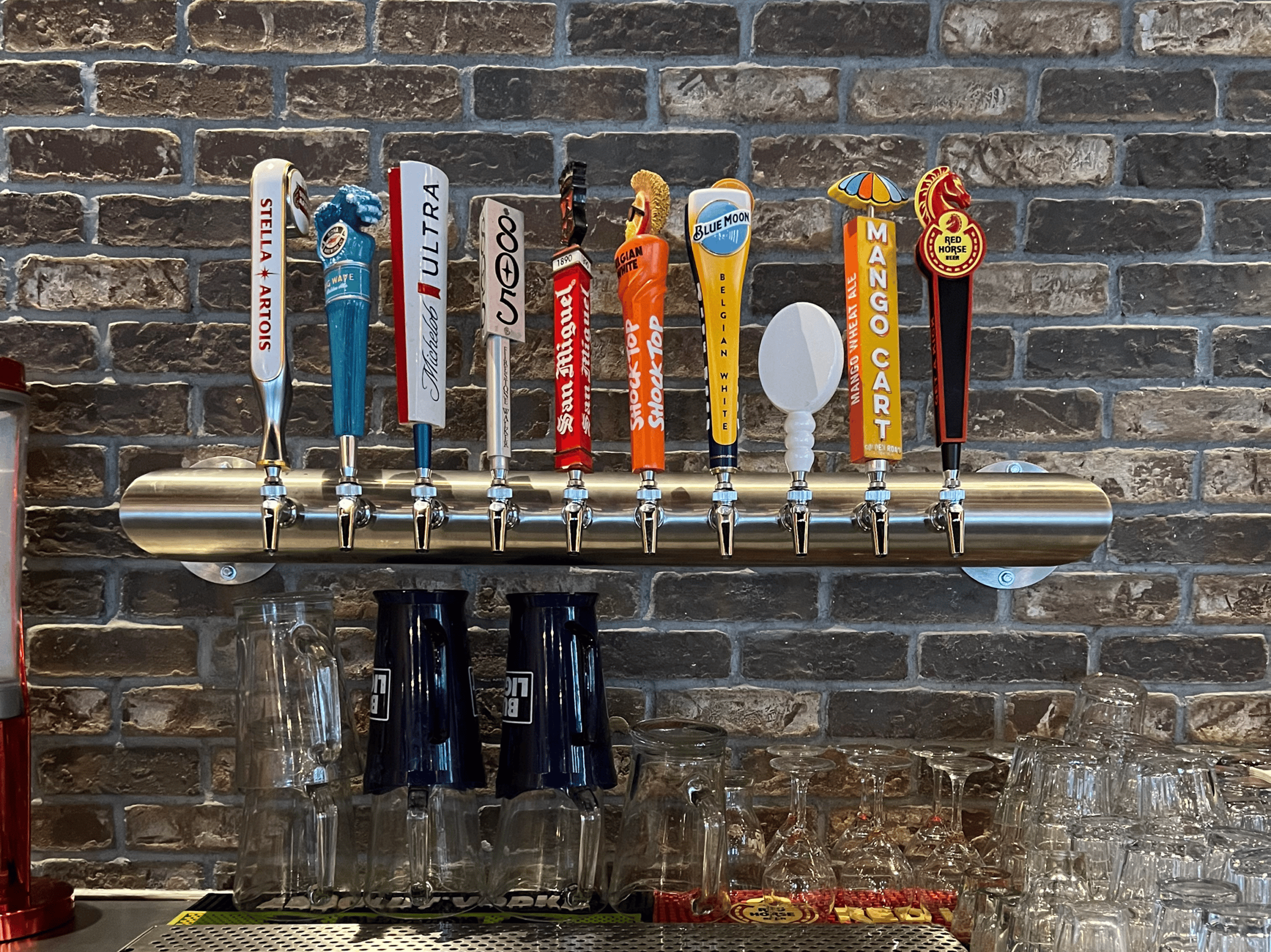 Multiple draft beer taps with colorful handles on a metal rail against a brick wall.